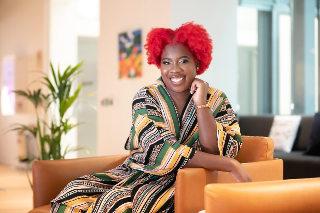 Dominique is a Caribbean woman with vibrant red curly. She is sitting on a brown leather sofa, wearing a colorful striped robe in a modern indoor space.